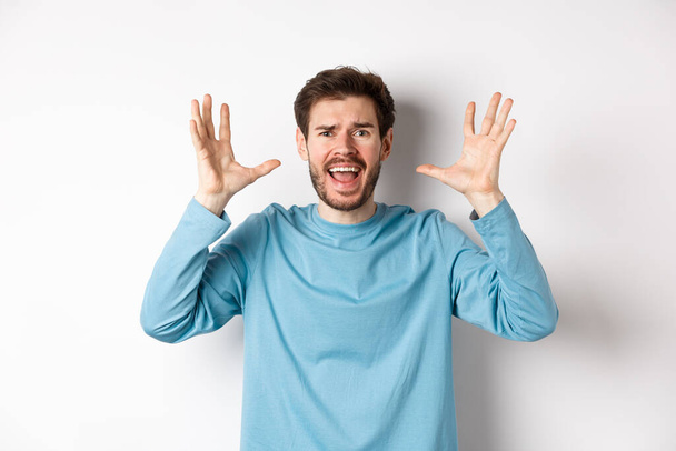 Image of freaked out young man screaming and shaking hands, shouting at camera frustrated or worried, panicking over white background - Photo, Image