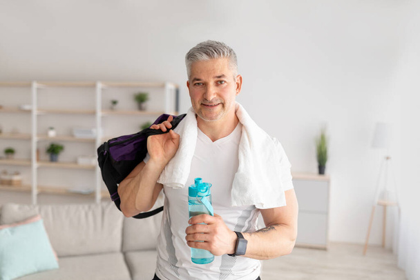 Motivation und sportlicher Lebensstil. Glücklicher reifer Mann mit Handtuch auf den Schultern, Wasserflasche und Sporttasche im Wohnzimmer - Foto, Bild