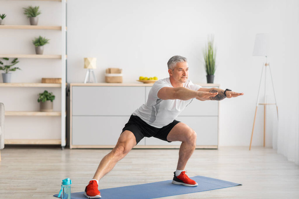 Home workout. Active mature man doing side lunges, stretching legs on yoga mat in living room, copy space - Photo, Image