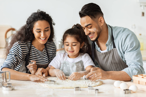 Θετικός άραβας πατέρας, μητέρα και κόρη που φτιάχνουν μπισκότα, φορώντας ποδιές - Φωτογραφία, εικόνα