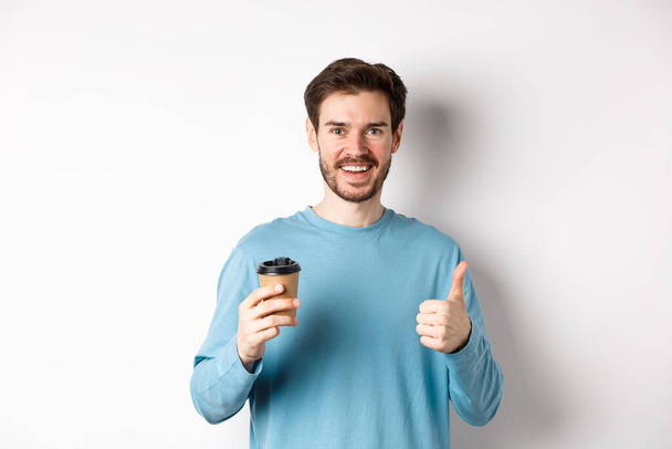 Χαρούμενος νεαρός άνδρας πίνοντας καλό καφέ, κρατώντας χάρτινο κύπελλο και δείχνοντας τον αντίχειρα επάνω, συνιστώντας cafe shop, στέκεται πάνω από το λευκό φόντο - Φωτογραφία, εικόνα
