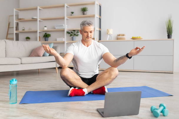 Dojrzały mężczyzna siedzi w pozycji lotosu i medytuje, siedzi na macie do jogi w salonie przed laptopem - Zdjęcie, obraz
