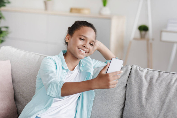 Joyeux adolescent afro américaine fille fait selfie pour les réseaux sociaux, assis sur le canapé à la maison dans le salon - Photo, image