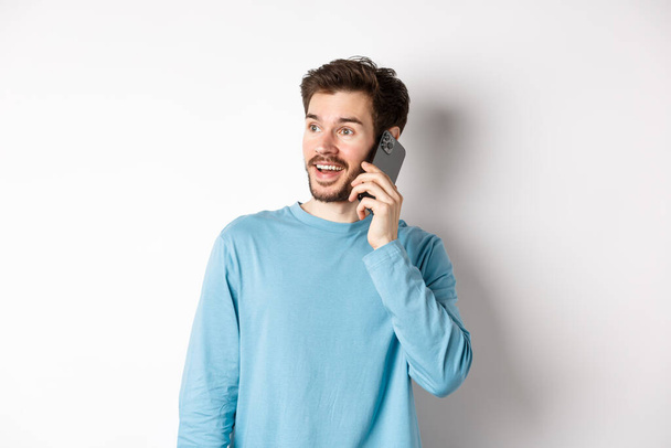 Concepto tecnológico. Hombre alegre hablando en el teléfono móvil, teniendo llamada telefónica y mirando divertido, de pie sobre fondo blanco - Foto, Imagen