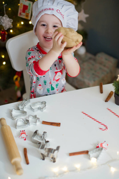 Menino bonito fazendo biscoitos de Natal em uma mesa branca perto da árvore de Natal com luzes. Conceito de cozinha de Natal
 - Foto, Imagem