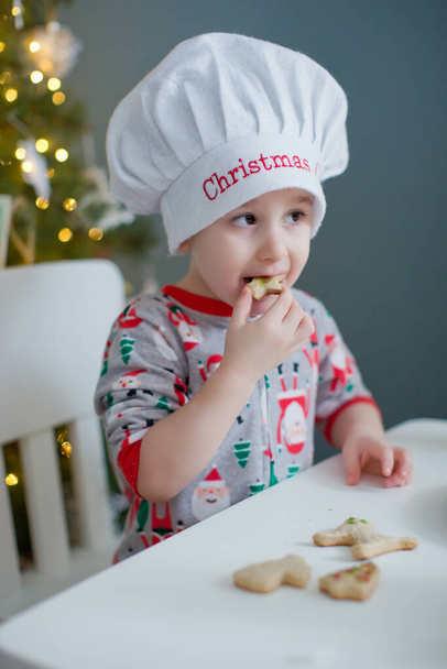 Söpö taapero poika syö joulukeksejä valkoisella pöydällä lähellä joulukuusi valot. Joulun ruoanlaitto käsite - Valokuva, kuva
