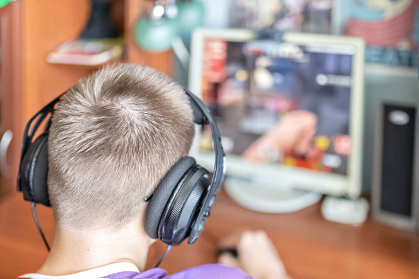 Teinipoika, nuori mies, joka pelaa videopelejä tietokoneella, käyttää teknologiaa, käyttää kuulokkeita, käyttää tietokonetta.Teini-ikäinen, joka käyttää teknologiaa. Hän pelaa videopelejä. Kuulokkeiden käyttö - Valokuva, kuva