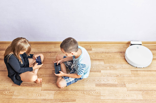 男の子と女の子の幼児が互いに反対側の床に座っています。彼らはスマートフォンでオンラインゲームをプレイします。Z世代,家族,電話,使用法,モバイル.トップ表示. - 写真・画像