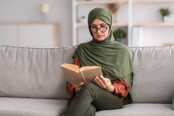 Ώριμο μουσουλμανικό γυναικείο βιβλίο ανάγνωσης φορώντας γυαλιά που κάθονται στο σπίτι - Φωτογραφία, εικόνα