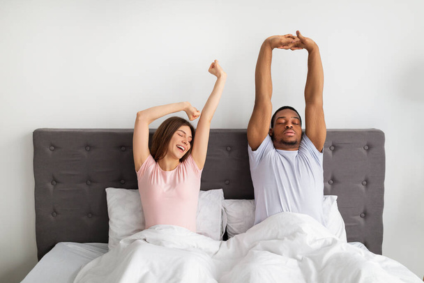 Πολυεθνικό ζευγάρι που τεντώνεται στο κρεβάτι μετά το ξύπνημα, νυστάζει στο σπίτι - Φωτογραφία, εικόνα