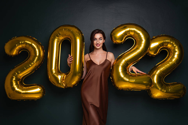 Happy young brunette woman in glamour dress near golden 2022 balloons on black background. New Year celebration. Smiling. Looking in the camera  - Fotoğraf, Görsel