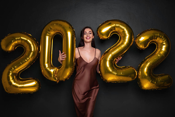 Happy young brunette woman in glamour dress near golden 2022 balloons on black background. New Year celebration. Smiling. Looking in the camera. Laughing. Holding numbers air balloons  - Valokuva, kuva