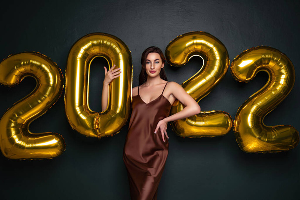 Happy young brunette woman in glamour dress near golden 2022 balloons on black background. New Year celebration. Looking in the camera. standing between the numbers air balloons  - Foto, Imagen
