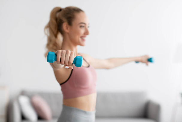Cheerful european millennial blonde female lifting dumbbells at home, selective focus - Photo, Image