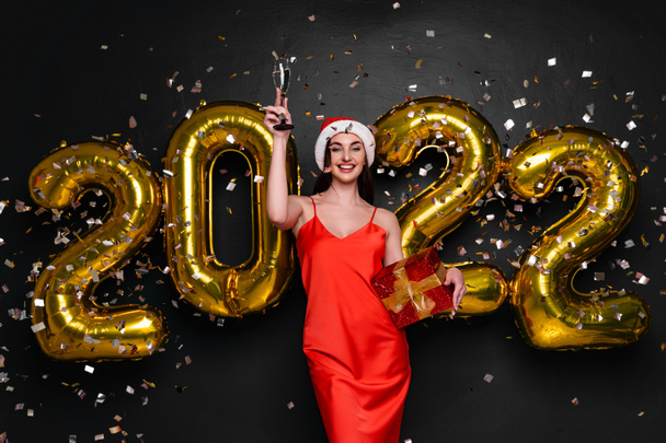 New Year Party. Happy smiling woman in santa hat and red dress raises the glass of champagne up. Celebrating holiday with gold 2022 balloons and confetti. Holding red gift box. Black background  - 写真・画像