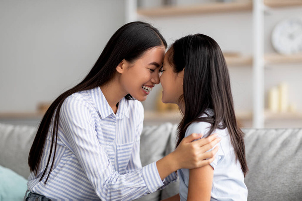 Schattig aziatisch moeder en dochter knuffelen en aanraken van elkaar met voorhoofd terwijl zitten op de bank in de woonkamer - Foto, afbeelding