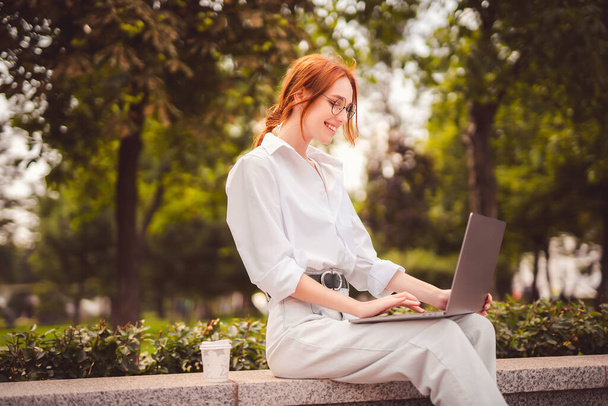 Beautiful redhead young woman sitting in the park and using laptop. Student. University. Freelance. Wearing in glasses jeans and white shirt. Coffee to go. Paper cup. Looking in laptop. Smiling  - Foto, immagini