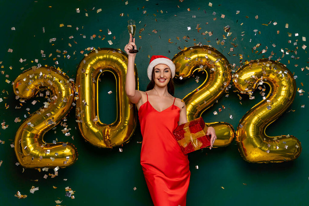 Happy smiling woman in santa hat and red dress raises the glass of champagne up. Celebrating holiday with gold 2022 balloons and confetti. Holding red gift box. Green background  - Foto, imagen