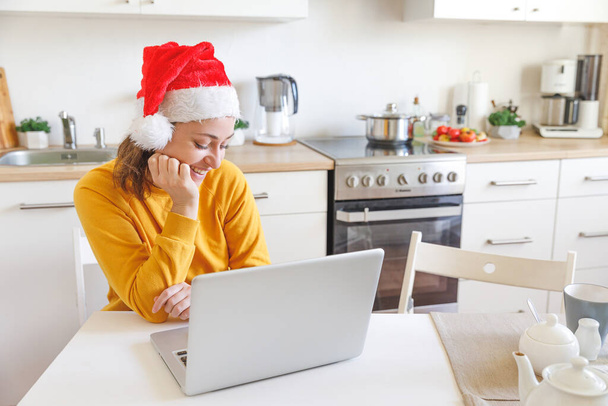 Usmívající se dívka v klobouku Santa mají video volá rodinu webkamerou. Žena s notebookem sedí doma v kuchyni a používá virtuální setkání chatu o prázdninách. Veselé Vánoce a Nový rok normální - Fotografie, Obrázek