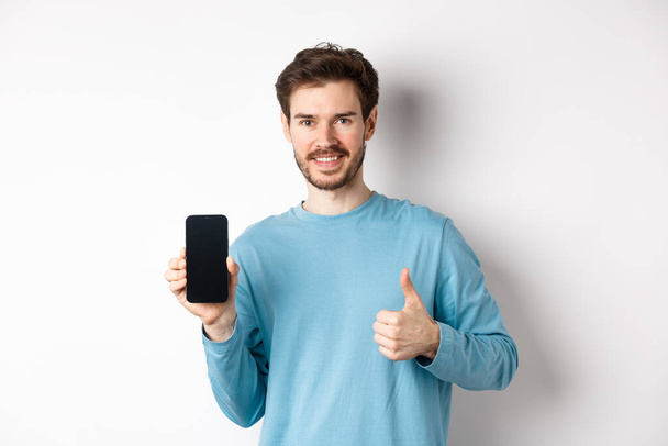 Giovane uomo sorridente mostrando schermo dello smartphone vuoto e pollici in su, lodando buona applicazione, raccomandare app mobile, in piedi su sfondo bianco - Foto, immagini