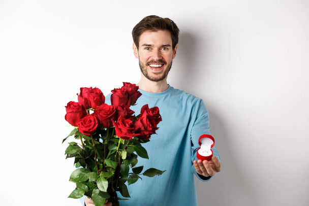 Όμορφος νεαρός άνδρας φίλος κάνει μια πρόταση για την ημέρα του Αγίου Βαλεντίνου εραστές, κρατώντας μπουκέτο από κόκκινα τριαντάφυλλα και δαχτυλίδι αρραβώνων, έννοια του γάμου και της σχέσης - Φωτογραφία, εικόνα