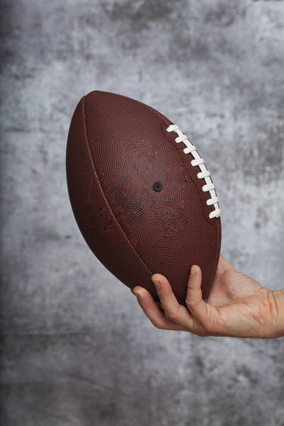 茶色のアメリカンフットボールボールを持っている男の右手テクスチャグレーの背景に白い文字列. - 写真・画像