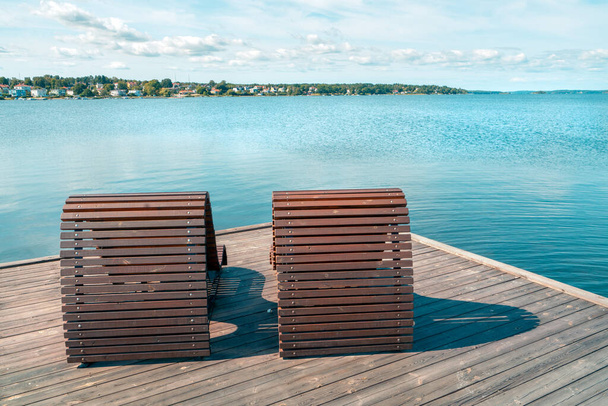 Két fából készült tengerparti ágy vagy szék egy fából készült mólón a tenger mellett, városképpel a háttérben. Napsütéses nyári nap a Balti-tenger mellett Vastervik, Dél-Svédország. Nyaralás a tengerparton. - Fotó, kép