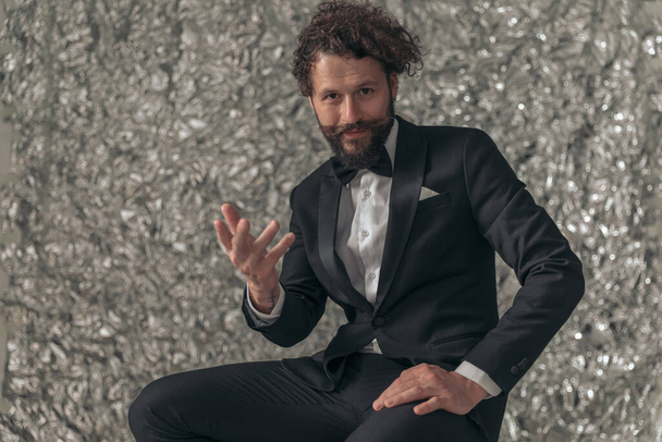 portrait of happy bearded elegant man in black tuxedo holding hand up, gesticulating and inviting while posing on tinfoil background in studio - Photo, Image