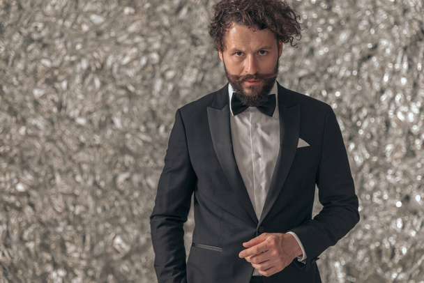 sexy young groom in black elegant tuxedo holding hands in pockets and posing in front of tinfoil background in studio - Φωτογραφία, εικόνα