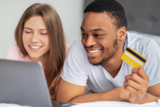 Porträt eines glücklichen multiethnischen Paares mit Kreditkarte und Laptop, das auf dem Bett liegt und von zu Hause aus online einkauft - Foto, Bild