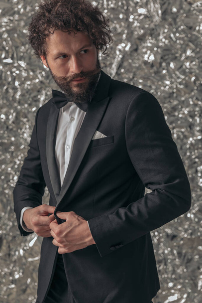 sexy bearded guy in elegant outfit looking to side and buttoning tuxedo while posing on tinfoil background in studio - Photo, Image