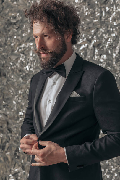 sexy elegant guy with long beard and moustache arranging tuxedo and looking to side on tinfoil background in studio - Photo, Image