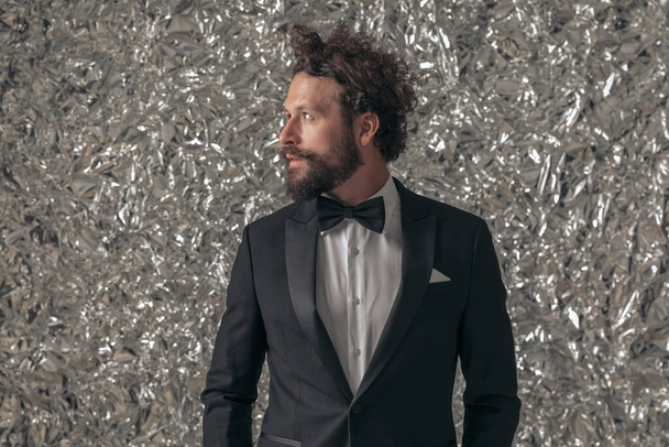 portrait of unshaved elegant groom in black tuxedo looking to side and holding hands in pockets while confidently posing in front of tinfoil background in studio - Photo, Image