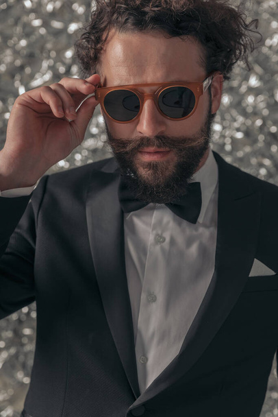 retrato de atractivo novio fresco en esmoquin negro ajustando gafas de sol y posando frente al fondo de papel de aluminio en el estudio - Foto, Imagen