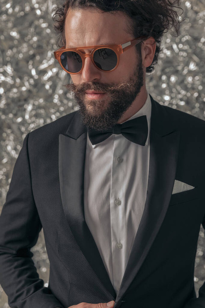 chladný elegantní muž v černém smokingu se slunečními brýlemi, který se dívá na stranu a upevňuje bundu, sebevědomě pózuje na alobalu pozadí ve studiu - Fotografie, Obrázek