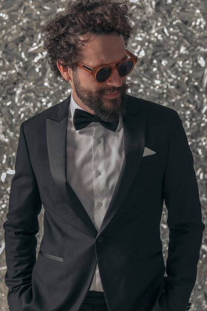 unshaved young groom in black tuxedo wearing sunglasses, holding hands in pockets and looking to side on tinfoil background in studio - Photo, Image