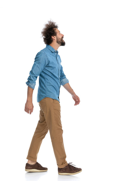 vista lateral de un hombre casual caminando en su camino y mirando hacia arriba sobre fondo blanco - Foto, Imagen