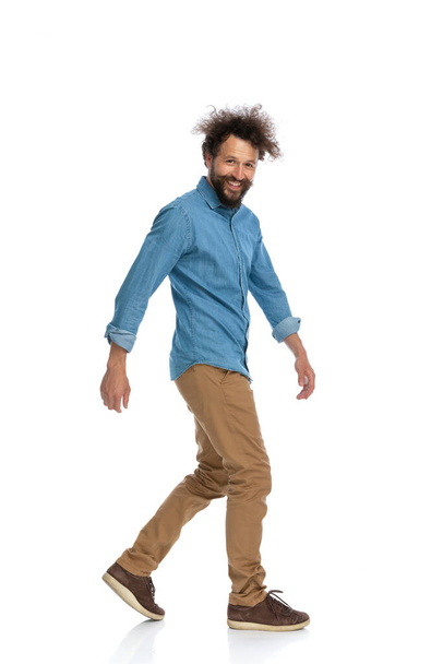 vista lateral de un hombre casual caminando hacia un lado y sonriendo a la cámara sobre fondo blanco - Foto, Imagen