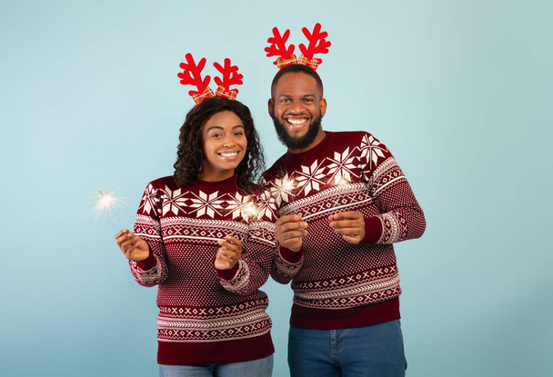 Šťastný africký americký pár v jelení rohy slaví Vánoce nebo Nový rok s jiskry přes modré pozadí - Fotografie, Obrázek