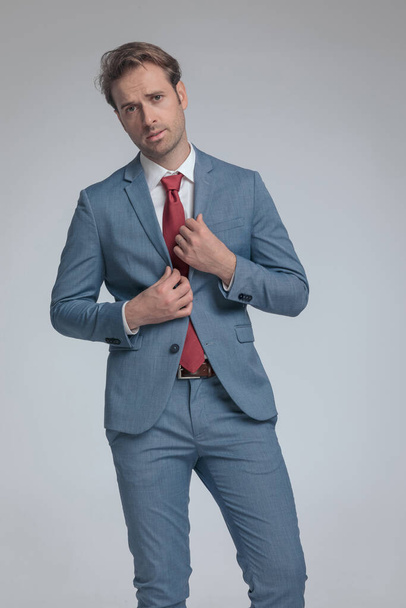 sexy Geschäftsmann arrangiert seinen Anzug und posiert mit Haltung auf grauem Hintergrund - Foto, Bild