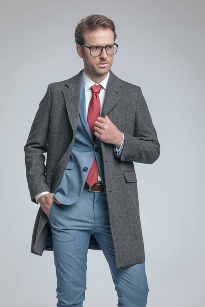 sexy fashion model sticking a hand in pocket, fixing his coat and wearing eyeglasses on gray background - Photo, Image