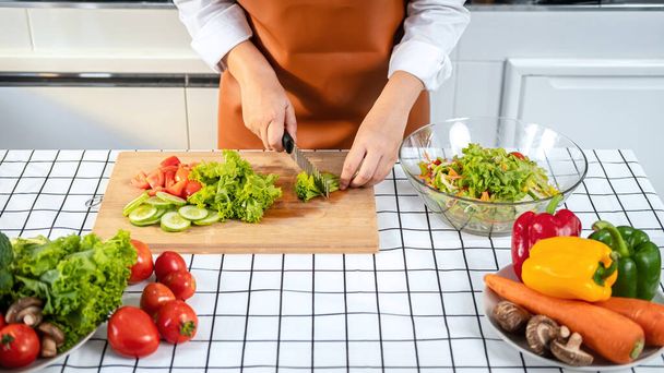 Asian housewife using knife to slice lettuce on wooden chopping board to preparing ingredients for vegetable salad while wearing apron and standing to cooking healthy meal in the kitchen at home - Photo, image