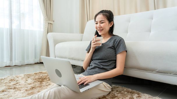 Азійська фрилансер жінка п'є чашку кави і використовує лептоп, щоб отримати електронну пошту замовлення від клієнта під час сидіння на підлозі, щоб перевірити продукт на фондовій біржі під час онлайн-маркетингової доставки в домашньому офісі. - Фото, зображення