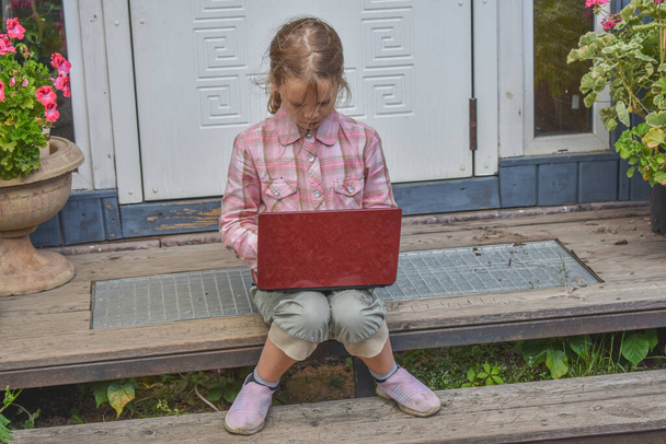 a girl child with a laptop is sitting on the porch of the house during a lesson on a remote photo without a filter - 写真・画像