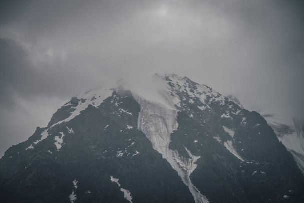 Dark atmospheric mountain landscape with glacier on black rocks in lead gray cloudy sky. Snowy mountains in low clouds in rainy weather. Gloomy landscape with black rocky mountains with snow in fog. - Photo, Image