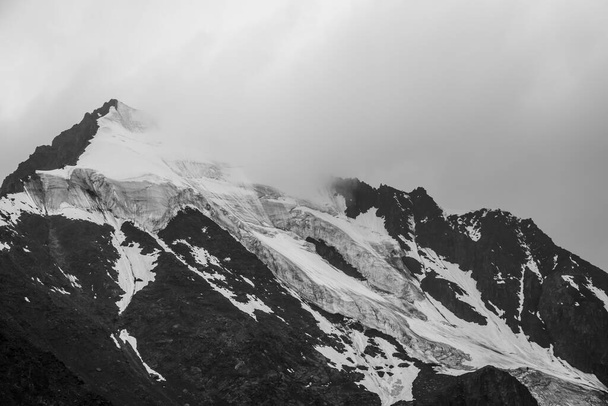 Monochrome atmospheric mountains landscape with big snowy mountain top in low clouds. Awesome minimal scenery with white glacier on black rocks. High mountain pinnacle with snow in clouds in grayscale - Photo, Image