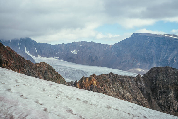 Awesome mountain scenery with layers of rocks and mountains with snow and glacier in high altitude under cloudy sky. Scenic view from snow cornice to glacier tongue among sharp rocks in low clouds. - Photo, Image
