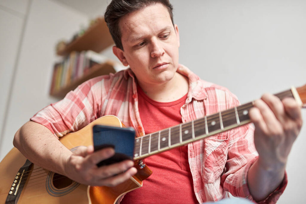 Junger Mann lernt, zu Hause mit Hilfe seines Smartphones Gitarre zu spielen. Konzepte virtueller Bildung, Hobbys und Musik. - Foto, Bild