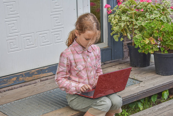 a girl child with a laptop is sitting on the porch of the house during a lesson on a remote photo without a filter - Zdjęcie, obraz