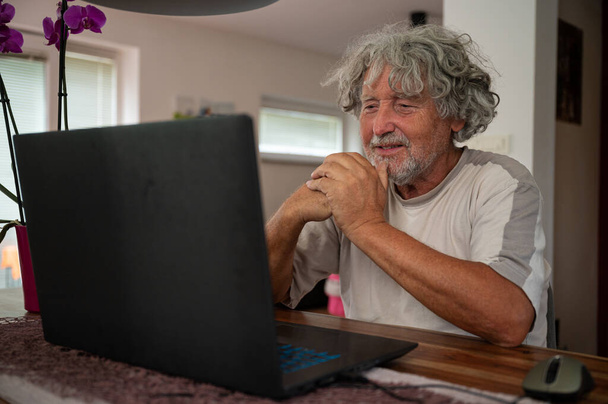 Старший человек, сидящий за обеденным столом с ноутбуком перед ним, разговаривает со своей семьей по видеосвязи. - Фото, изображение